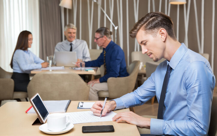 Serious young businessman writing at table in cafe with blurred business team sitting at table with laptop and working in background. Side view.