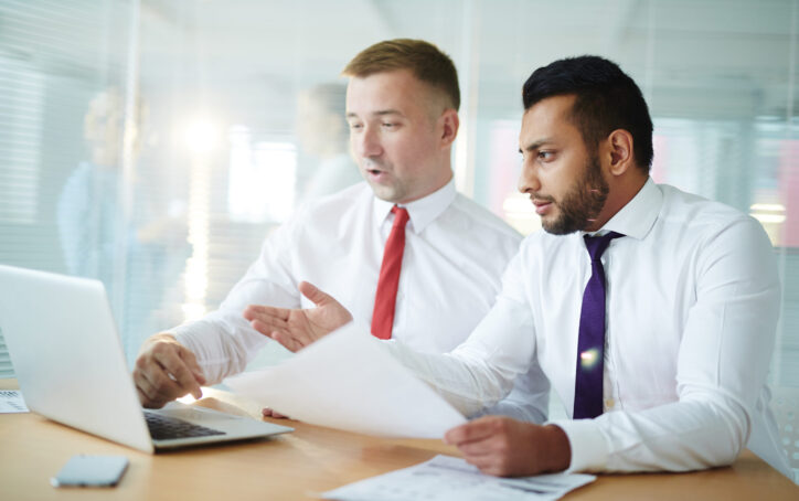 Two businessmen sitting in front of laptop and looking through online financial statistics in office