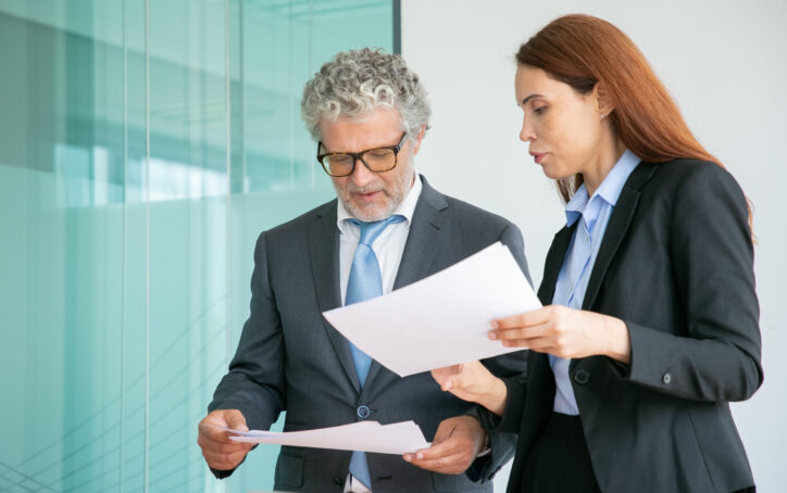 Professional partners discussing project and holding papers in conference room. Successful concentrated grey-haired entrepreneur talking, standing and reading. Business and management concept
